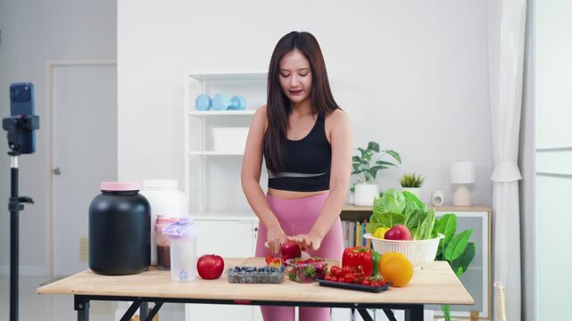 Wellness influencer talking about weight loss diet while filming in modern kitchen. Beautiful Asian woman producing engaging video content about natural healthy eating habits.