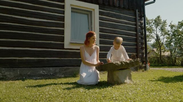 A woman and her little boy sit on a stone outside a traditional house in Bucovina. They laugh and enjoy their time together in the sunny weather.