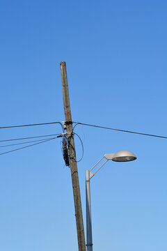 An old wooden utility pole carrying telecommunication cables stands beside a modern metal street light under a clear blue sky.