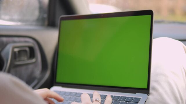 Person types on laptop with green screen inside car. Fingers move over keyboard, focused on work. Scene shows modern, mobile productivity concept. Useful for advertising remote work, tech gadgets