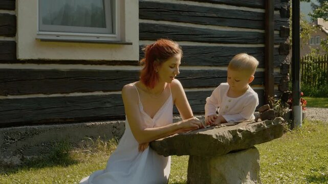 A woman and her little boy enjoy their time outside near a traditional house in Bucovina. They play with stones and share smiles in the warm sun.