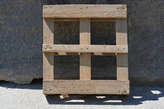 A square wooden industrial shipping pallet stands upright leaning against a gray textured concrete wall on an asphalt ground.