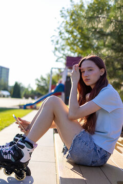 Teenage girl wearing roller skates sits outdoors and uses a smartphone while taking break after skating on warm summer evening. Concept of digital lifestyle, Gen Z and mobile communication.