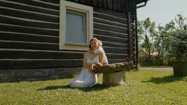 A woman sits outside a traditional house in Bucovina while her little boy hugs her. They share a moment of joy in the sunny weather, surrounded by nature.