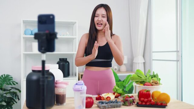 Wellness influencer talking about weight loss diet while filming in modern kitchen. Beautiful Asian woman producing engaging video content about natural healthy eating habits.