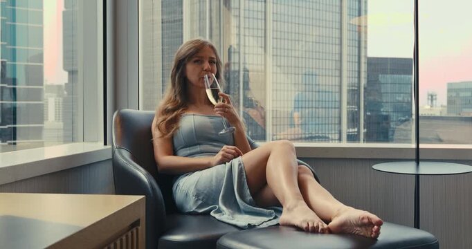 Woman relaxes in a leather armchair sipping champagne at a luxury hotel room in Hong Kong during spring afternoon, shot as an eye-level medium shot.
