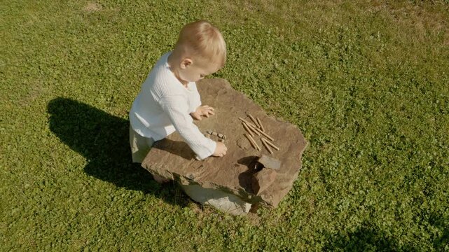 A little boy spends his time outside a traditional house playing with pebbles and pencils on a rock. He enjoys his simple activities in the sun.