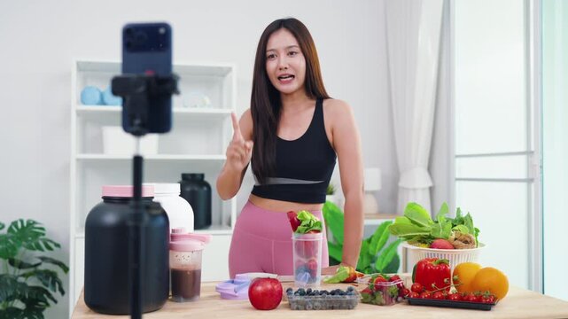 Wellness influencer talking about weight loss diet while filming in modern kitchen. Beautiful Asian woman producing engaging video content about natural healthy eating habits.
