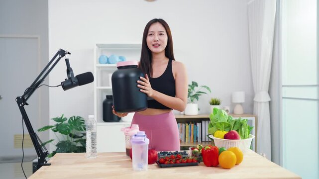 Young female vlogger making diet coaching video with fruits and blender in kitchen. Asian sporty woman streaming live tutorial about organic meals and holistic wellbeing.