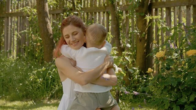 A woman and her little boy play together in their yard. They smile and hug each other while enjoying their time among the flowers in the sunny afternoon.