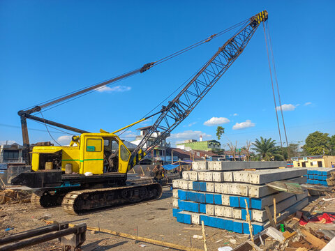 Yellow crawler crane with lattice boom on a construction site next to stacks of precast concrete piles.