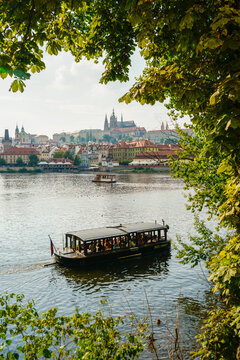 Barco tur&iacute;stico navegando por el r&iacute;o Moldava con vistas al castillo de Praga y el centro hist&oacute;rico rodeado de vegetaci&oacute;n verde.