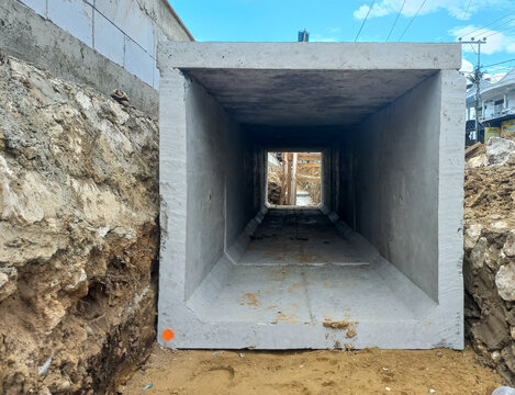 Precast concrete box culvert installed in an excavated trench for drainage and infrastructure. The large square concrete tunnel sits between earthen walls with blue sky above.