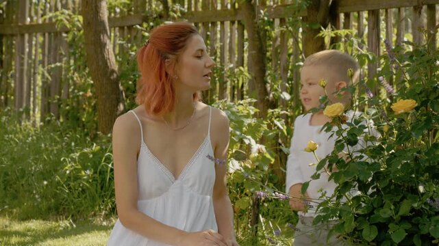 A woman sits in the yard while her little boy explores the flowers. They share a fun moment outside, enjoying the warmth of the sun and nature around them.
