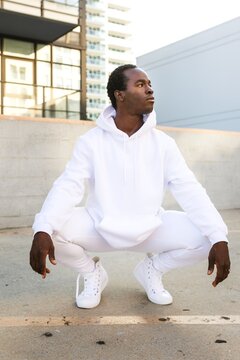 Person in white hoodie and sneakers squatting outdoors, urban setting.