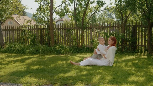 A woman sits on the grass while her little boy runs to her. They hug and roll around. This family time happens in a sunny yard with trees around.