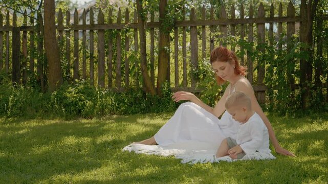 A woman sits on the grass with her little boy as they explore their yard together. They have fun playing and learning about the outdoors on a sunny day.