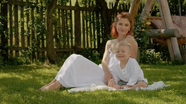 A woman and her son sit on the grass in their yard, looking at the camera. They enjoy a sunny day together, creating family memories while relaxing outdoors.