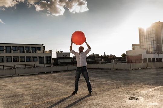 Mature man playing with orange fitness ball on rooftop of a high-rise building