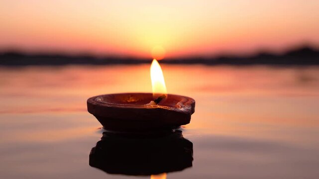 A traditional clay lamp diya with a flickering flame floats on tranquil water at sunset, reflecting the warm glow. The scene evokes peace and spiritual reflection.