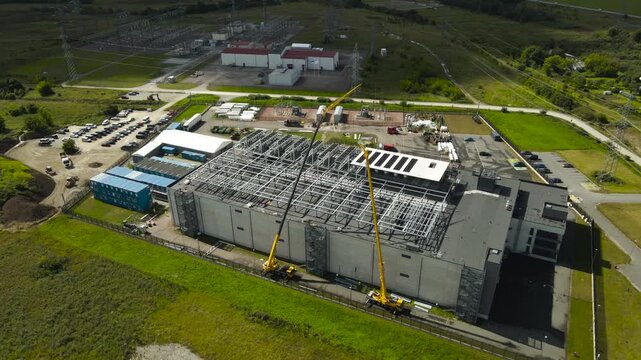 High angle view showing two large mobile cranes lifting and positioning steel trusses on the roof of a modern data center building, with an electrical substation visible in the background