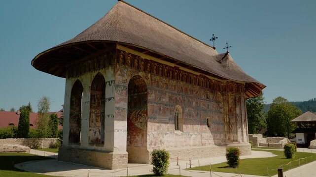 Visitors walk around Humor Monastery in Bucovina, Romania. The site shows beautiful old paintings on the walls and a unique structure. It offers a look into history.