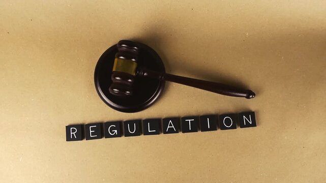 Top down view of a wooden judge's gavel and sound block beside the word regulation, spelled out in black letter tiles on a brown paper background, symbolizing law, order, and authority