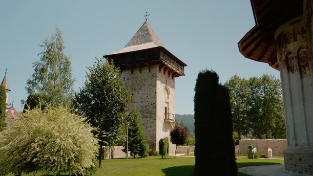 Humor monastery shows old walls and a tall tower under a clear sky. The area has green trees and gardens all around. People can walk and enjoy the space.