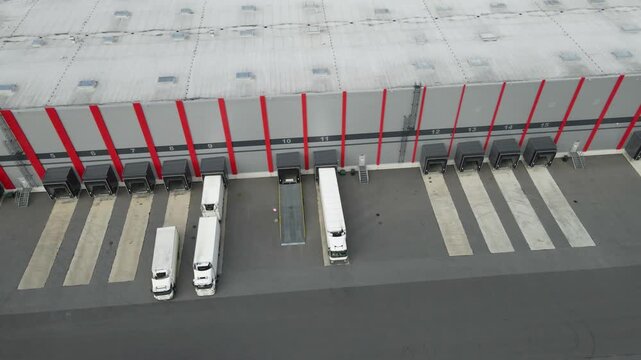 High angle view showing a large semi trailer truck backing into a loading bay at a modern logistics center. The industrial building features multiple ramps for cargo and freight operations