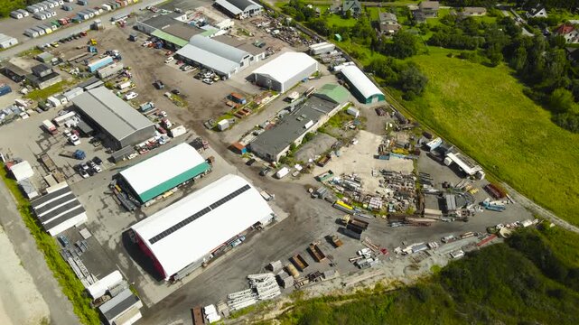 High altitude perspective flying over a large scrapyard filled with piles of junk, old vehicles, and scrap metal next to industrial buildings and warehouses in an urban area during the day