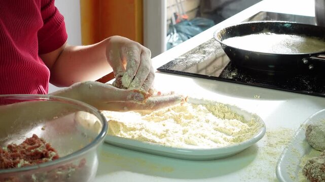 Girl hands shaping and flouring homemade raw beef meatballs on a white tray on the kitchen counter. Next to her a hot pan ready to fry them
