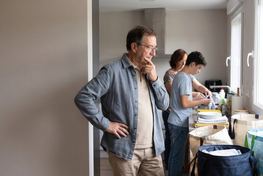 Grandfather contemplating while woman and boy unpack essentials and clean in kitchen