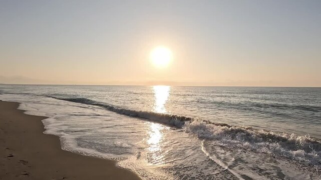 Slow motion video walking along the brown sand of the beach shore watching the sun at sunrise or sunset over the horizon of the calm Mediterranean Sea, with a clear sky
