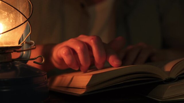 Female student studying and reading textbook by candlelight of vintage oil lamp while sitting at desk in dark room at home in evening