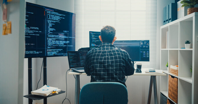 Young Caucasian male programmer writes code using multiple screens in home office workspace. AI development, cybersecurity, network database system, software engineering, remote tech startup concept.
