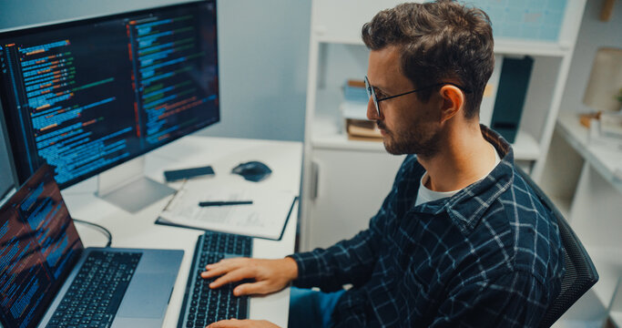 Close up young Caucasian male programmer writes code using multiple screens in home office workspace. AI development, cybersecurity, network database system, software engineering, remote tech startup.