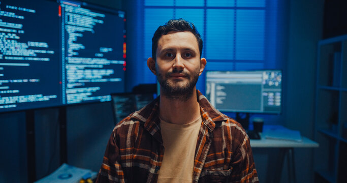 Portrait of confident Caucasian male programmer smiles looking at camera in office at night. Professional developer workspace, Secure computer network system, Advanced cybersecurity data concept.