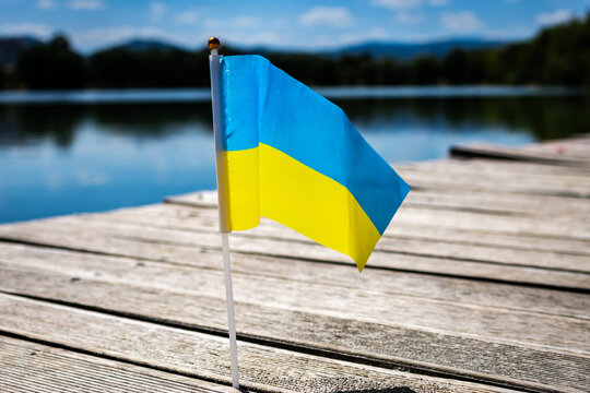 Small Ukrainian flag on a wooden pier beside a calm lake under a bright blue sky. A light breeze lifts the blue and yellow fabric. Peaceful summer scene with copy space.