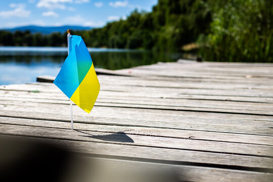 Mini Ukrainian flag planted in pier boards beside a tranquil lake. Bright blue and yellow cloth ripples in warm sunshine with distant hills and soft bokeh giving room for copy.