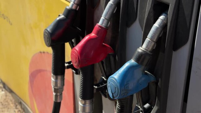 Fuel dispensers display red and blue nozzles being removed from their holders at a gas station, with a bright outdoor environment visible in the background