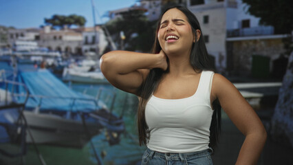 Fototapeta premium Woman with hand on neck wincing for neck pain in white tank top and jeans in studio with marina backdrop; discomfort strain.