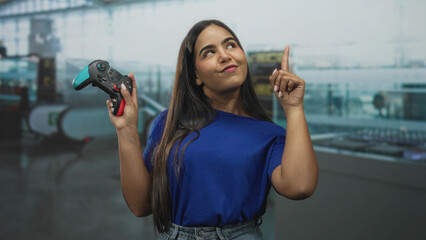 Woman in blue shirt holds gamepad with raised hand and points finger up in airport terminal by escalator  joy confidence gaming success. © Krakenimages.com