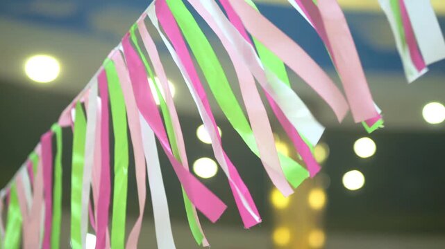 Colorful ribbons dangle in a festive setting. Pink green and white strips flutter gently. Warm lights create a soft glow in the background. This decoration evokes joyful celebrations