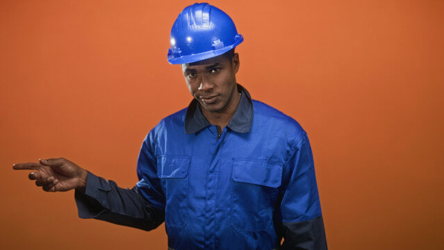 Man in blue hardhat and blue coveralls points finger to left, visible hand and finger, wearing safety helmet and work uniform in studio; focus guidance.