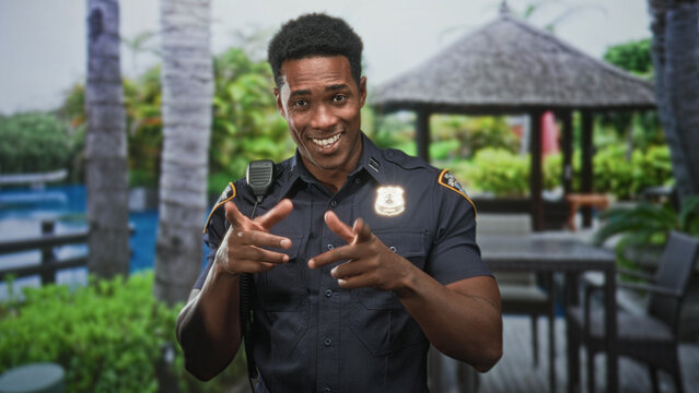 Man police officer in uniform with badge and radio mic pumping right fist while touching chest near building gazebo; confidence celebration.