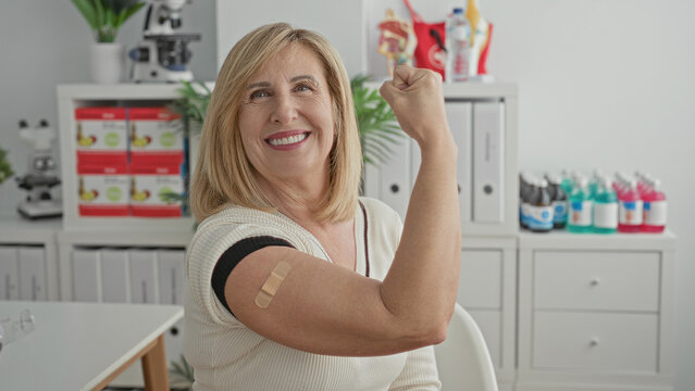 Woman flexes arm with bandage in clinic, smiling while seated and showing vaccination site with raised fist and visible skin; confidence wellness recovery.