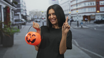 Fototapeta premium Young woman smiling, holding orange pumpkin bucket with finger pointing up on a street; playful celebration.