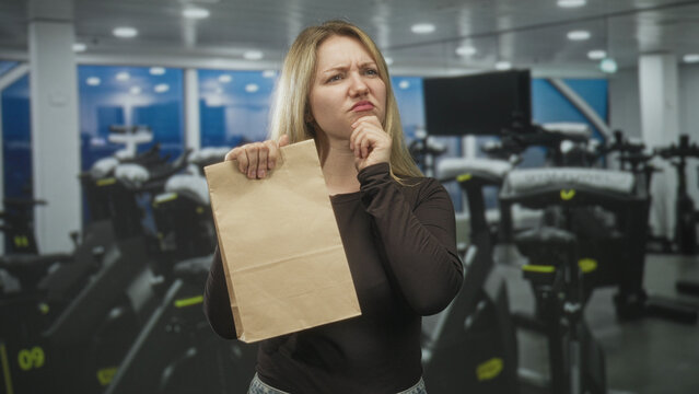 Woman holds paper bag and touches chin in spin studio while frowning and posing; skepticism thinking.