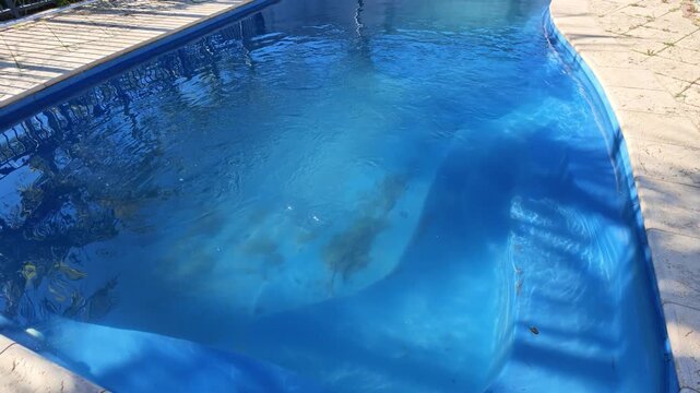 Dirty swimming pool with sand and algae at the bottom needing maintenance. The rippling blue water reflects the sunny day and surroundings