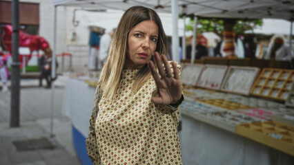 Fototapeta premium Woman with palm raised in a stop gesture by a street market stall, patterned blouse and rings visible, direct serious gaze; defiance.
